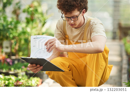 Notepad in hands. Young man with curly hair and in glasses is in greenhouse 132203051