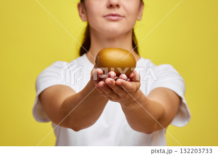 Happy woman in white t-shirt holding kiwi on studio yellow background, focus on kiwi 132203733