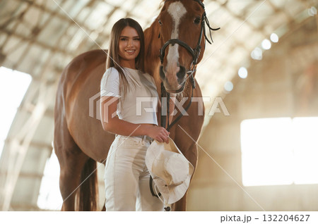 Holding cowboy hat in hand. Beautiful young woman is with horse indoors 132204627
