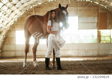 Holding cowboy hat in hand. Beautiful young woman is with horse indoors 132204629
