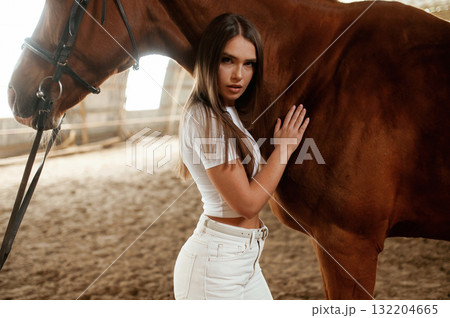 Touching animal by hand and looking at the camera. Beautiful young woman is with horse indoors Touching animal by hand and looking at the camera. Beautiful young woman is with horse indoors 132204665