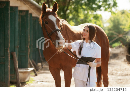 Holding notepad. Female vet is with horse outdoors at the farm at daytime Holding notepad. Female vet is with horse outdoors at the farm at daytime 132204713