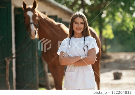 Arms crossed, standing and smiling. Female vet is with horse outdoors at the farm at daytime 132204832