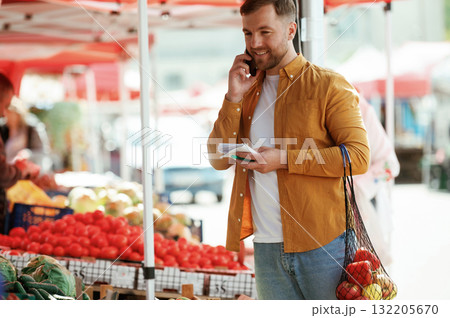 Talking by phone. With shopping list. Handsome man is on the street market or bazaar Talking by phone. With shopping list. Handsome man is on the street market or bazaar 132205670