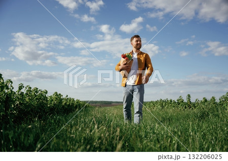 Shopping bag with products in hands. Handsome man is on the agricultural field at daytime Shopping bag with products in hands. Handsome man is on the agricultural field at daytime 132206025