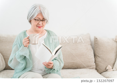 ソファーでホットドリンクを飲みながら読書するシニアのアジア人女性 ソファーでホットドリンクを飲みながら読書するシニアのアジア人女性 132207097