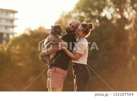 Lovely couple, holding animal in hands. Man and woman are with dog on the field Lovely couple, holding animal in hands. Man and woman are with dog on the field 132207504