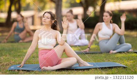 Sporty young girls performing seated twisting asana Matsyendrasana on mat in in summer park during daily yoga session Sporty young girls performing seated twisting asana Matsyendrasana on mat in in summer park during daily yoga session 132211120