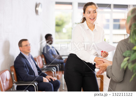 Smiling young girl makes deal, shaking hands with elderly woman colleague while standing in office. 132211122