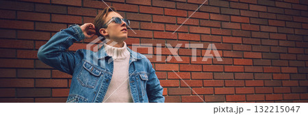 Confident man in denim jacket and sunglasses posing against brick wall in urban street. Banner Confident man in denim jacket and sunglasses posing against brick wall in urban street. Banner 132215047
