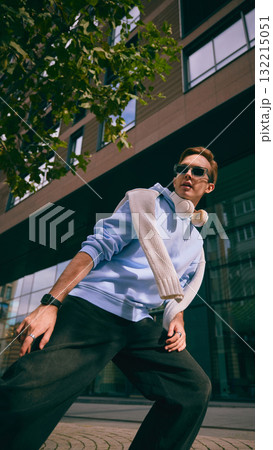Man in blue hoodie with headphones walking near glass building and trees 132215051