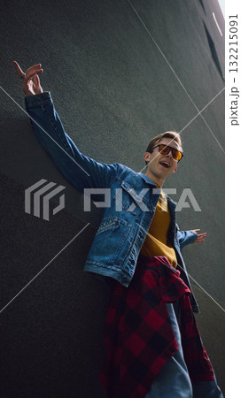 Man in denim jacket posing with arms wide open against tall gray wall 132215091