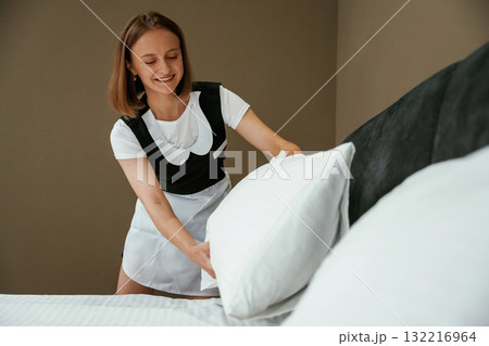 In uniform, taking care of bed. Young chambermaid is in hotel room 132216964