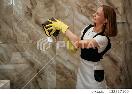 Glass in the bathroom is getting cleaned. Young chambermaid is in hotel room 132217109