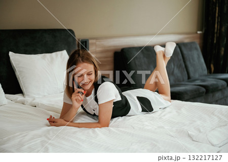 Cheerful facial expression, lying down on bed with phone. Young chambermaid is in hotel room Cheerful facial expression, lying down on bed with phone. Young chambermaid is in hotel room 132217127