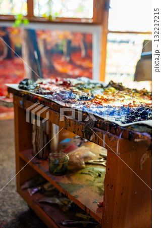 Artist creative workspace with oil paint palette, colorful brushstrokes, paint tubes, glass jar, chair and canvas in the background. Real studio atmosphere full of artistic energy. Artist creative workspace with oil paint palette, colorful brushstrokes, paint tubes, glass jar, chair and canvas in the background. Real studio atmosphere full of artistic energy. 132217215