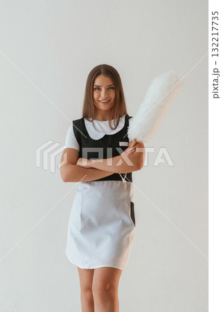 Positive, chambermaid is holding cleaning thing. Young pretty woman is against background in the studio 132217735