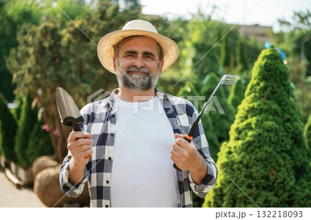 Standing with tools. Senior gardener is at work at daytime Standing with tools. Senior gardener is at work at daytime 132218093