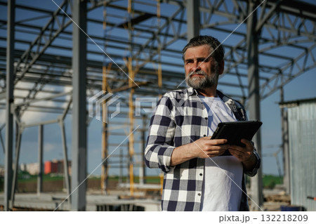 Wireless device, holding digital tablet in hands. Senior worker is near the unfinished greenhouse, working 132218209