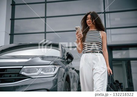 With smartphone. Attractive young woman is outdoors near her brand new car With smartphone. Attractive young woman is outdoors near her brand new car 132218322