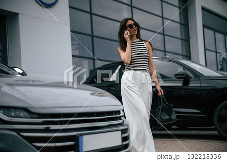 Talking by phone. Attractive young woman is outdoors near her brand new car Talking by phone. Attractive young woman is outdoors near her brand new car 132218336