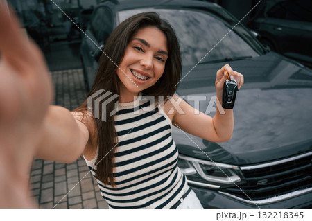 Making selfie, holding keys, happy owner. Attractive young woman is outdoors near her brand new car Making selfie, holding keys, happy owner. Attractive young woman is outdoors near her brand new car 132218345