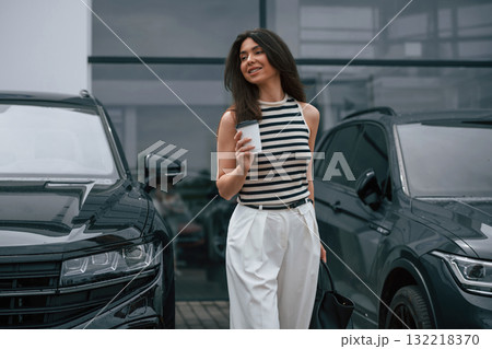 Walking holding drink. Attractive young woman is outdoors near her brand new car Walking holding drink. Attractive young woman is outdoors near her brand new car 132218370