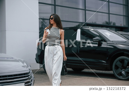 Walking holding drink. Attractive young woman is outdoors near her brand new car Walking holding drink. Attractive young woman is outdoors near her brand new car 132218372