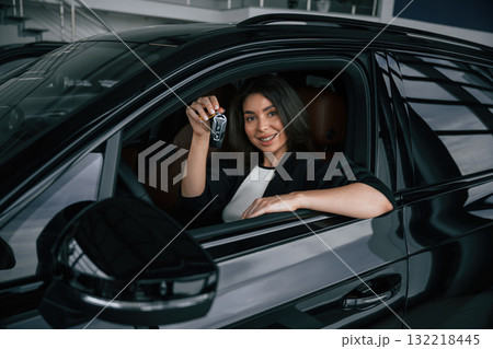 Looking out through the opened window. Happy woman is sitting in the new car and holding keys Looking out through the opened window. Happy woman is sitting in the new car and holding keys 132218445