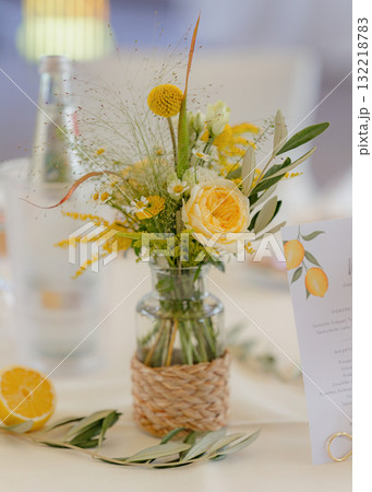 Floral arrangement with yellow and white flowers in a glass jar on a wedding table. Fresh lemon half, greenery, and menu card with a lemon design create a charming, rustic atmosphere Floral arrangement with yellow and white flowers in a glass jar on a wedding table. Fresh lemon half, greenery, and menu card with a lemon design create a charming, rustic atmosphere 132218783