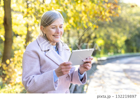 Senior woman in a sunny autumn park smiling as she uses a tablet, enjoying modern technology, relaxation and connection among golden leaves and natural light 132219897