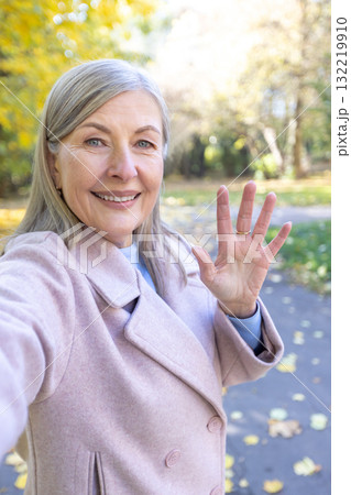 Senior woman in a stylish coat capturing a selfie while smiling and waving to the camera, enjoying the beautiful autumn day in the park with golden foliage 132219910