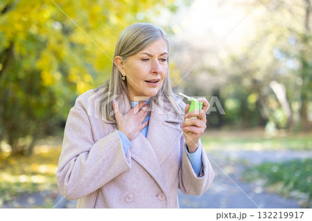 Senior gray-haired woman suffering from sore throat, using spray medicine to relieve pain. Woman walking in park. 132219917