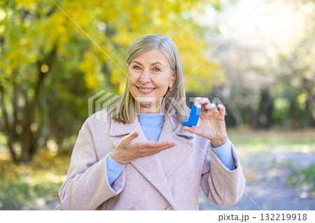 Senior woman smiling while holding a blue asthma inhaler in an autumn park, promoting respiratory health, confidence, vitality and outdoor wellbeing among older adults 132219918