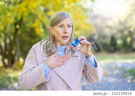 Female senior with an anxious expression experiencing an asthma attack in a park, holding an inhaler and placing a hand on her chest, seeking relief from breathing problems Female senior with an anxious expression experiencing an asthma attack in a park, holding an inhaler and placing a hand on her chest, seeking relief from breathing problems 132219919