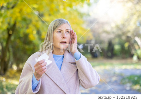 Senior woman feeling unwell with a headache, touching her temple and holding a tissue while standing outdoors in an autumn park, experiencing sickness and discomfort Senior woman feeling unwell with a headache, touching her temple and holding a tissue while standing outdoors in an autumn park, experiencing sickness and discomfort 132219925