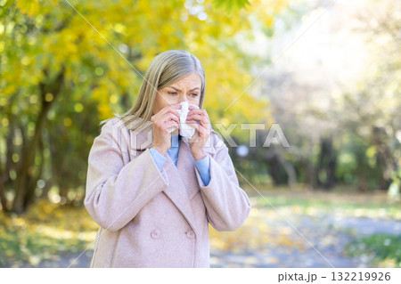 Senior woman blowing her nose into a tissue in an autumn park, battling allergy or cold symptoms amid yellow foliage, bundled in a coat and looking unwell Senior woman blowing her nose into a tissue in an autumn park, battling allergy or cold symptoms amid yellow foliage, bundled in a coat and looking unwell 132219926