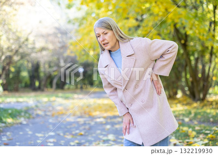 Senior woman clutching lower back in pain while walking through an autumn park, grimacing from chronic back ache and stiffness, posture bent from discomfort and mobility issues Senior woman clutching lower back in pain while walking through an autumn park, grimacing from chronic back ache and stiffness, posture bent from discomfort and mobility issues 132219930