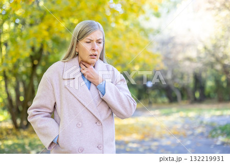 Senior woman standing in an autumn park, suffering from a sore throat and discomfort during the fall season, feeling unwell with hands touching her neck Senior woman standing in an autumn park, suffering from a sore throat and discomfort during the fall season, feeling unwell with hands touching her neck 132219931