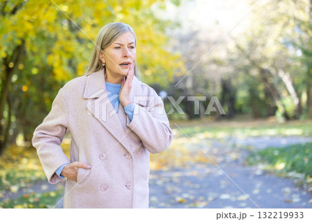Senior woman standing in an autumn park suffering from a toothache, holding hand to her cheek, expressing discomfort and dental pain, highlighting common health issues as people age 132219933