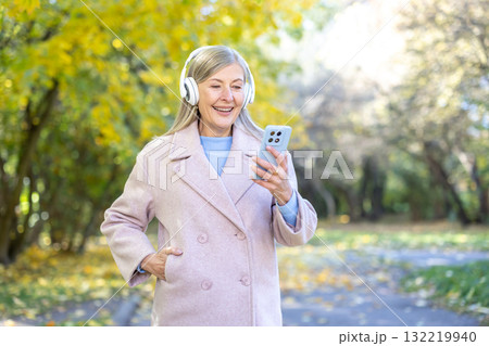 Senior woman strolling through an autumn park with headphones, smiling at her smartphone, enjoying music and nature on a bright, relaxed outdoor walk Senior woman strolling through an autumn park with headphones, smiling at her smartphone, enjoying music and nature on a bright, relaxed outdoor walk 132219940