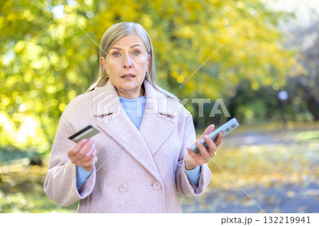 Confused senior woman in an autumn park holding a credit card and smartphone, shocked to discover an online scam or failed transaction, worried about fraud and finances Confused senior woman in an autumn park holding a credit card and smartphone, shocked to discover an online scam or failed transaction, worried about fraud and finances 132219941