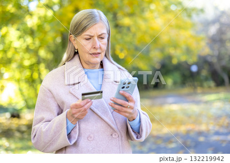 Senior woman outdoors looks puzzled holding credit card and smartphone, worried about online payment problems, potential scam or banking error while trying mobile transactions 132219942