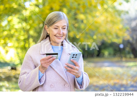 Senior woman smiling while holding a credit card and smartphone, making an online purchase or payment in a park during autumn, embracing modern technology 132219944