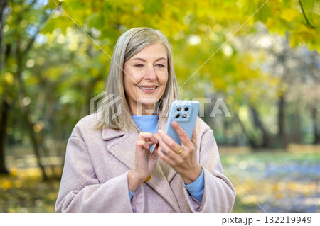 Senior woman smiling cheerfully while engaging with her smartphone outdoors, connecting with technology and enjoying social media in a beautiful autumn park environment 132219949