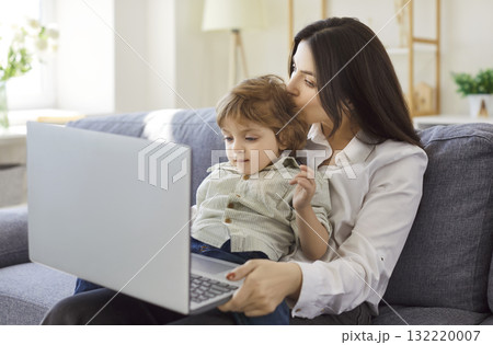 Mother working on laptop while cuddling toddler on sofa, balancing work and family Mother working on laptop while cuddling toddler on sofa, balancing work and family 132220007