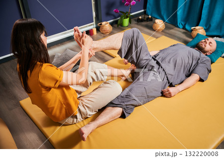 Masseuse making thai yoga massage. Woman therapist doing traditional massage treatment, stretching male patient's body. Client lying on mat, enjoying hand massage in calm atmosphere. 132220008