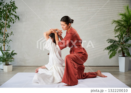 Masseuse making thai yoga massage. Woman therapist doing traditional massage treatment, stretching female patient's body. Client sitting on mat, enjoying hand massage in calm atmosphere. 132220010