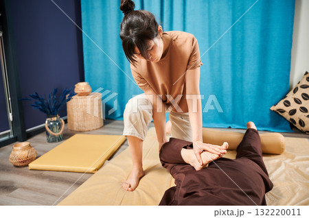 Masseuse making thai yoga massage. Woman therapist doing traditional massage treatment, stretching female patient's body. Client lying on mat, enjoying hand massage in calm atmosphere. Masseuse making thai yoga massage. Woman therapist doing traditional massage treatment, stretching female patient's body. Client lying on mat, enjoying hand massage in calm atmosphere. 132220011