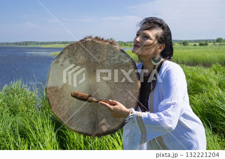 Female shaman in the white dress drumming outdoors 132221204